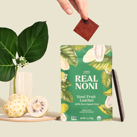 Noni Fruit Leather package with hand taking a serving piece displayed next to fresh ripe hawaiian noni fruit and noni leaves